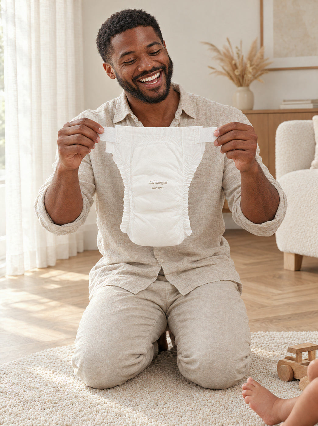 A smiling dad kneeling on a rug and holding up a fresh diaper by its tabs, printed dad changed this one