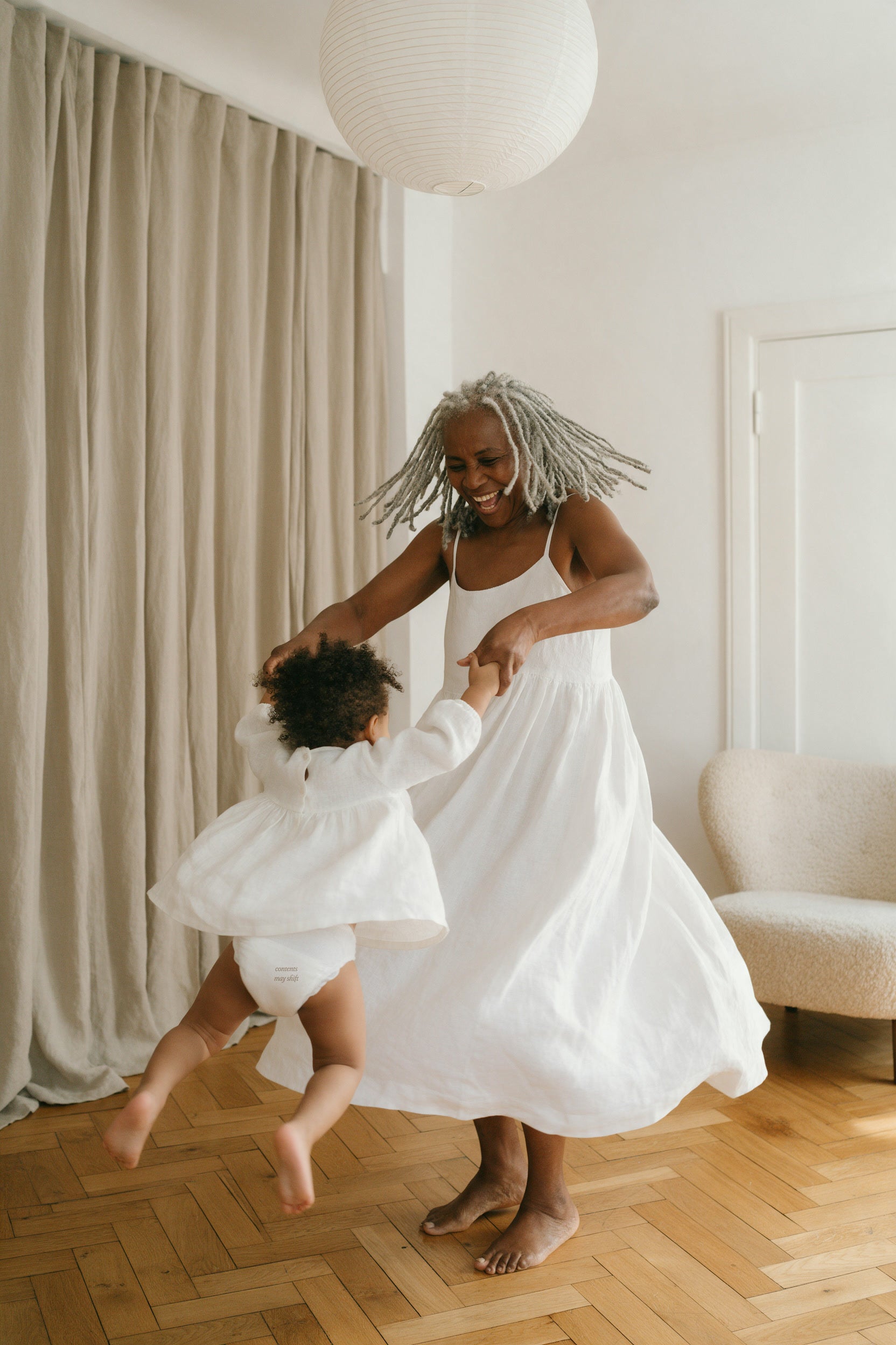 A grandmother in a white dress spinning a laughing toddler around a sunlit room, diaper printed contents may shift