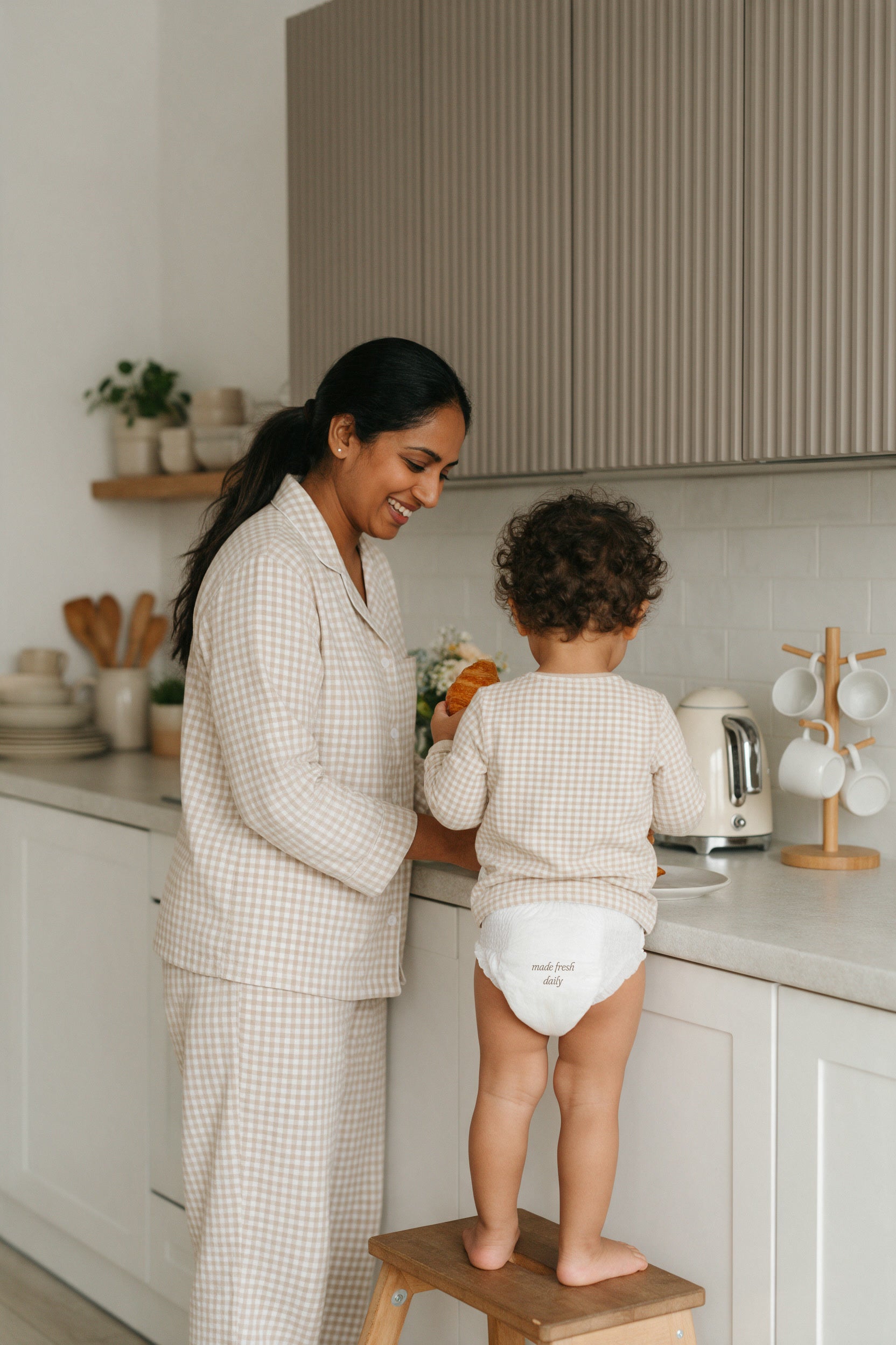 A toddler on a wooden step stool at the kitchen counter beside mom in gingham pajamas, diaper printed made fresh daily